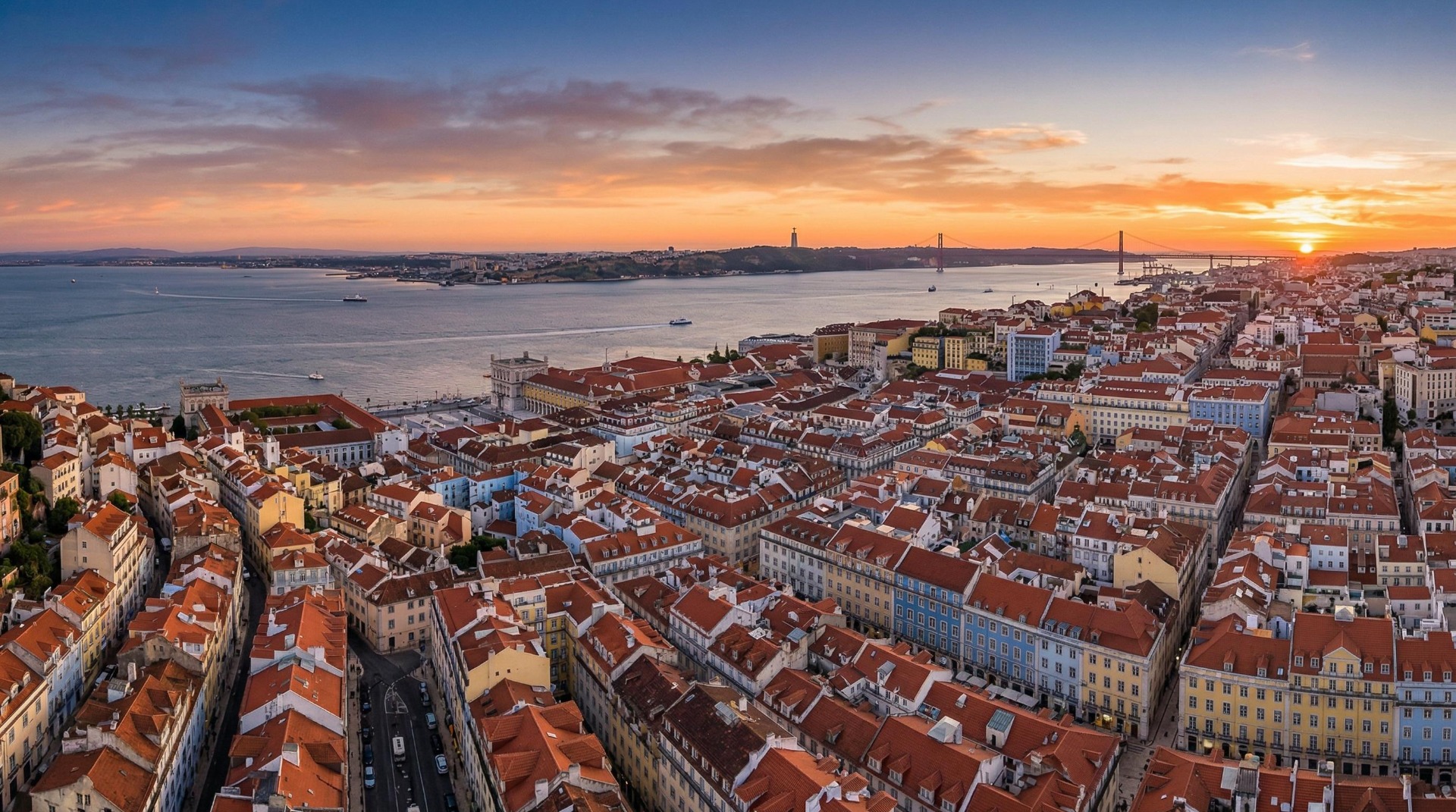 Vista aérea de telhados vermelhos e rio ao pôr do sol, com cidade costeira e barcos.